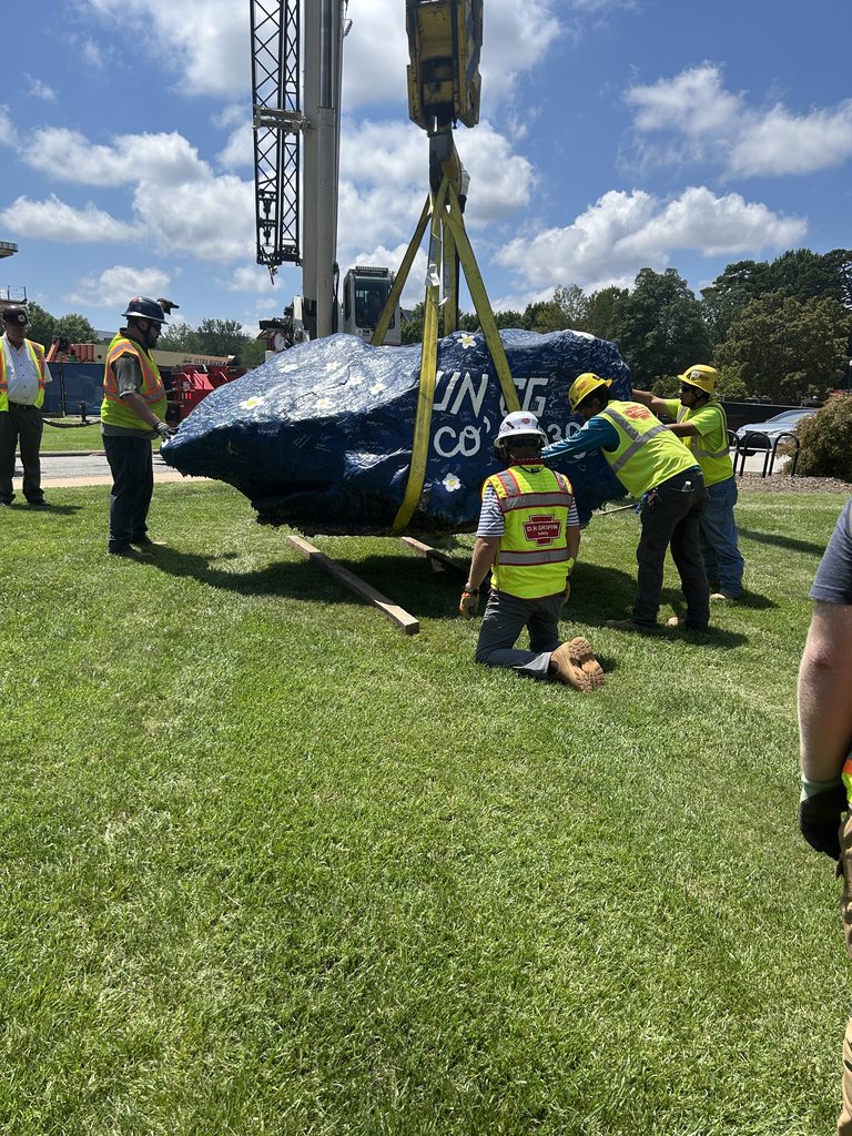 People standing and kneeling next to a large boulder which has been strapped to the end of a crane.