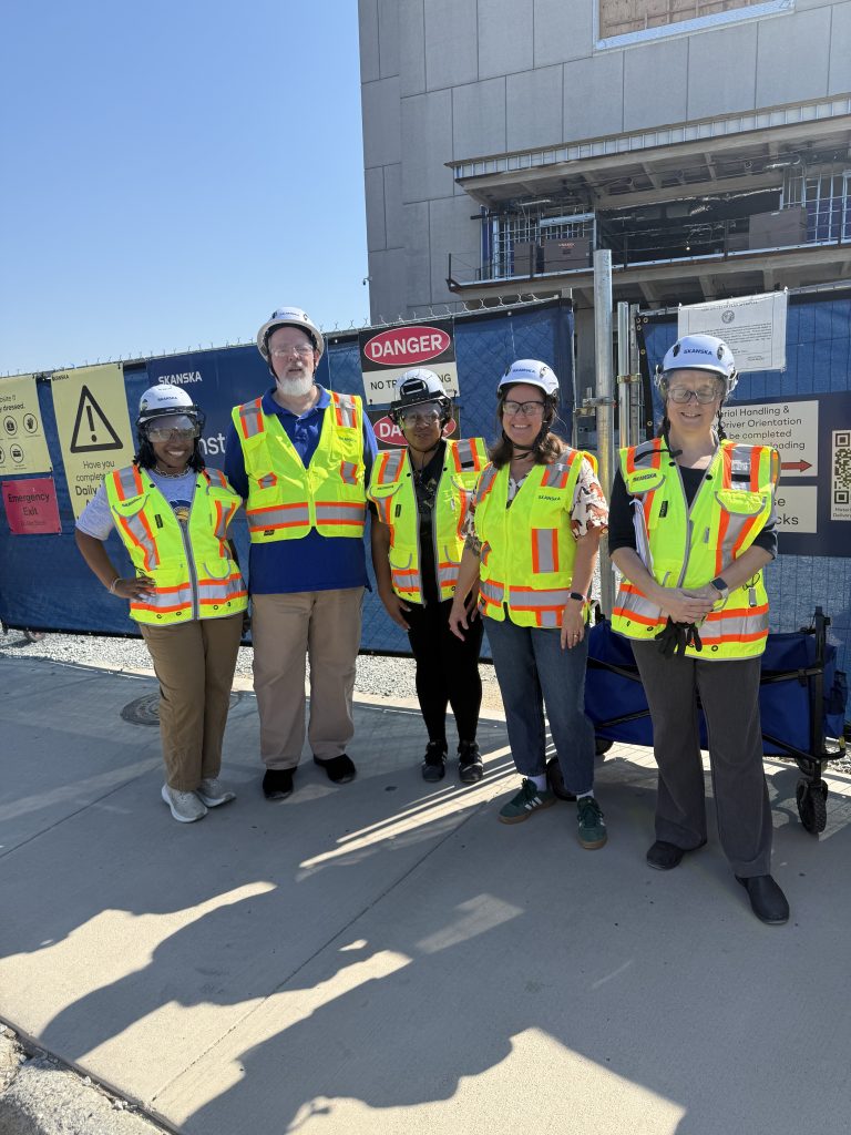 Five people standing next to one another in front of construction fencing. They are wearing protective vets, hats, and eyewear.